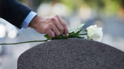 a man's hand lays a white rose on a curved grave stone.