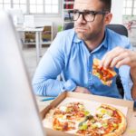 A sad man looks at his computer screen as he holds a slice of pizza in his hand with an open pizza box in front of him on his desk.