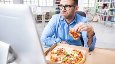 A man looks sadly away from his computer screen as he holds a slice of pizza in his hand with an open pizza box in front of him on his desk.
