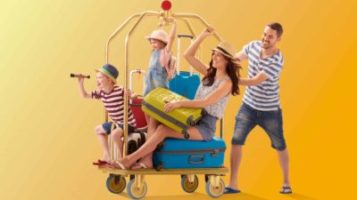 a family of parents with two children ride an airport trolley with luggage and tourist trappings such as field glasses with excited expressions on their faces.