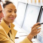 A woman points with her pen at a computer where a colleague sits as though they are collaborating on a project. She has a smile on her face.