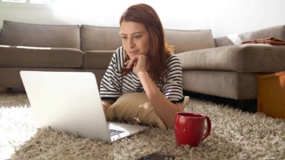 A smiling woman with a satisfied look on her face lies on a rug in her home with her laptop open and a large cup on the floor nearby, gazing at the screen. researching new ETFs