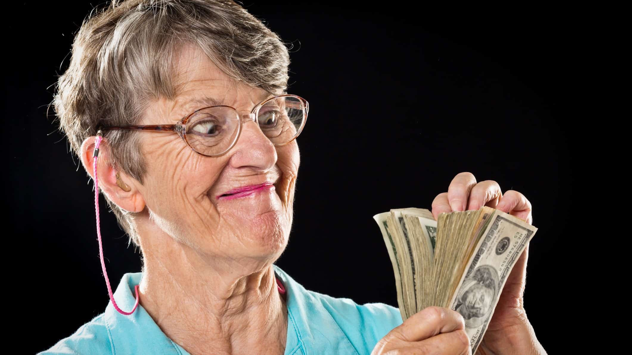 an older woman holds a handful of paper money in her hands and looks at them with a slightly crazy smile on her face wearing her spectacles on a string as a lot of older people do.