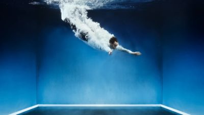 a man in business attire plunges into a room filled with water with bubbles streaming along his body as though he has completed a high dive.