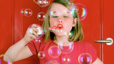a girl in a red t-shirt stands against a red door blowing bubbles through a red bubbleblower.