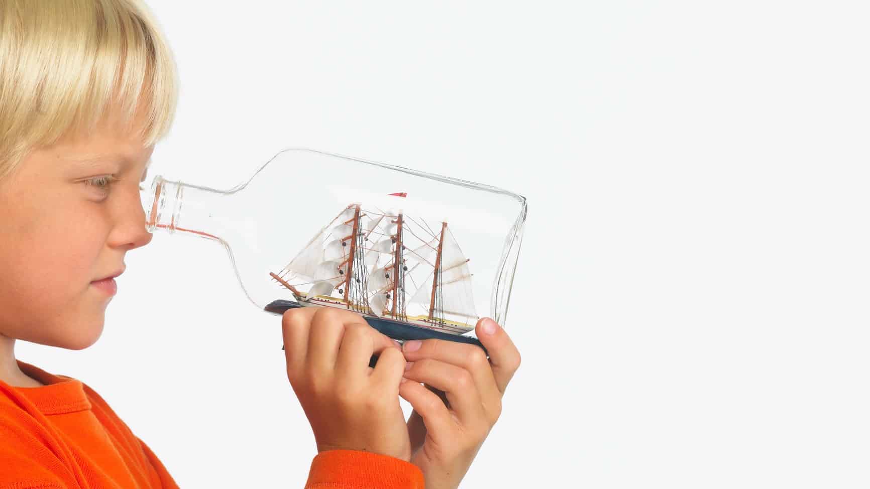 a boy looks into the top of a glass bottle that holds a model ship inside it.