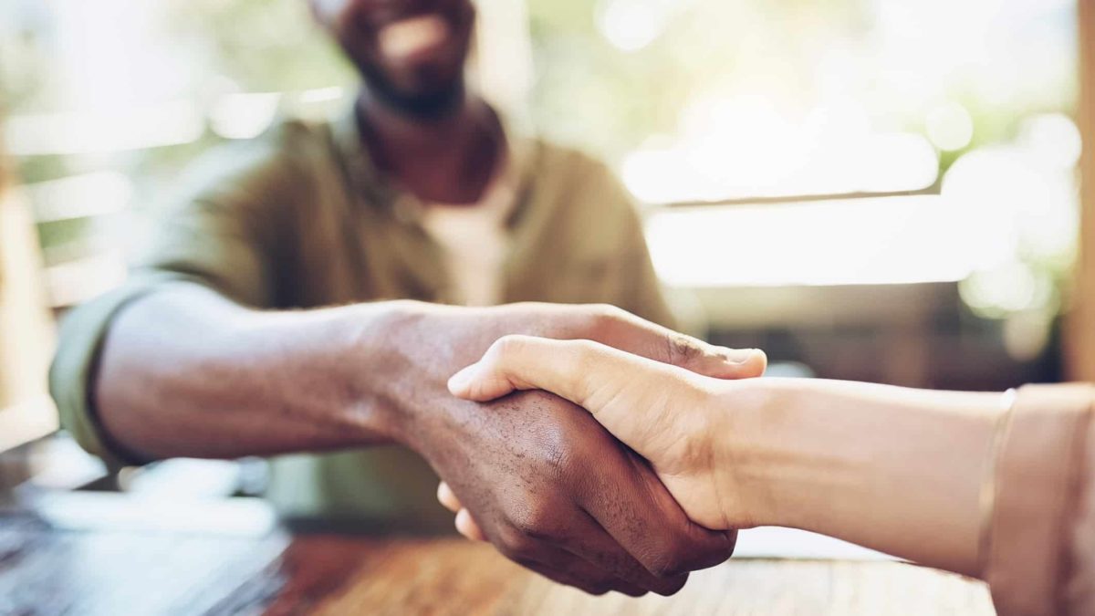 a close up of a happy handshake between two people, one of whom is grinning widely in the background, as though a beneficial deal has been struck between the two people.