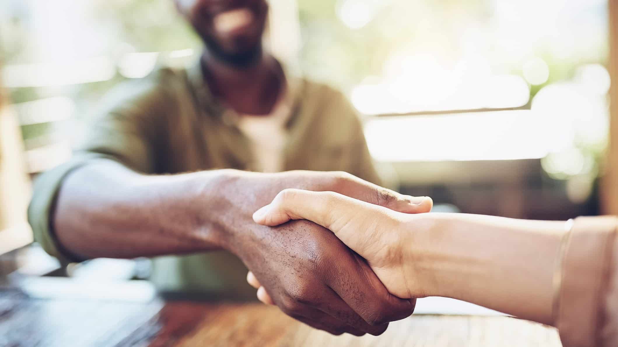 a close up of a happy handshake between two people, one of whom is grinning widely in the background, as though a beneficial deal has been struck between the two people.
