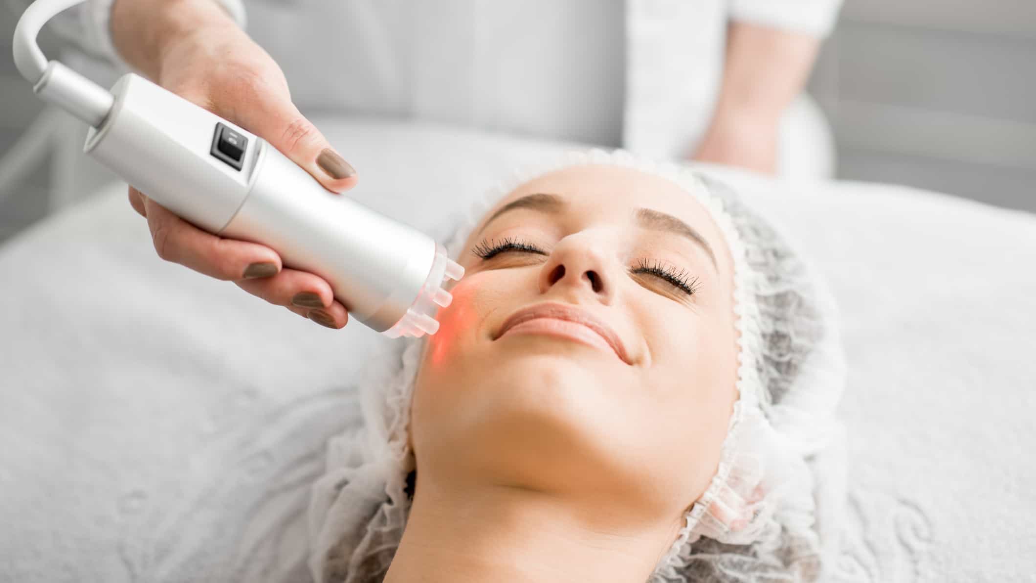 a woman lies on a medical bed for a cosmetic lasering session with a hand held laser directing light onto her cheek area.