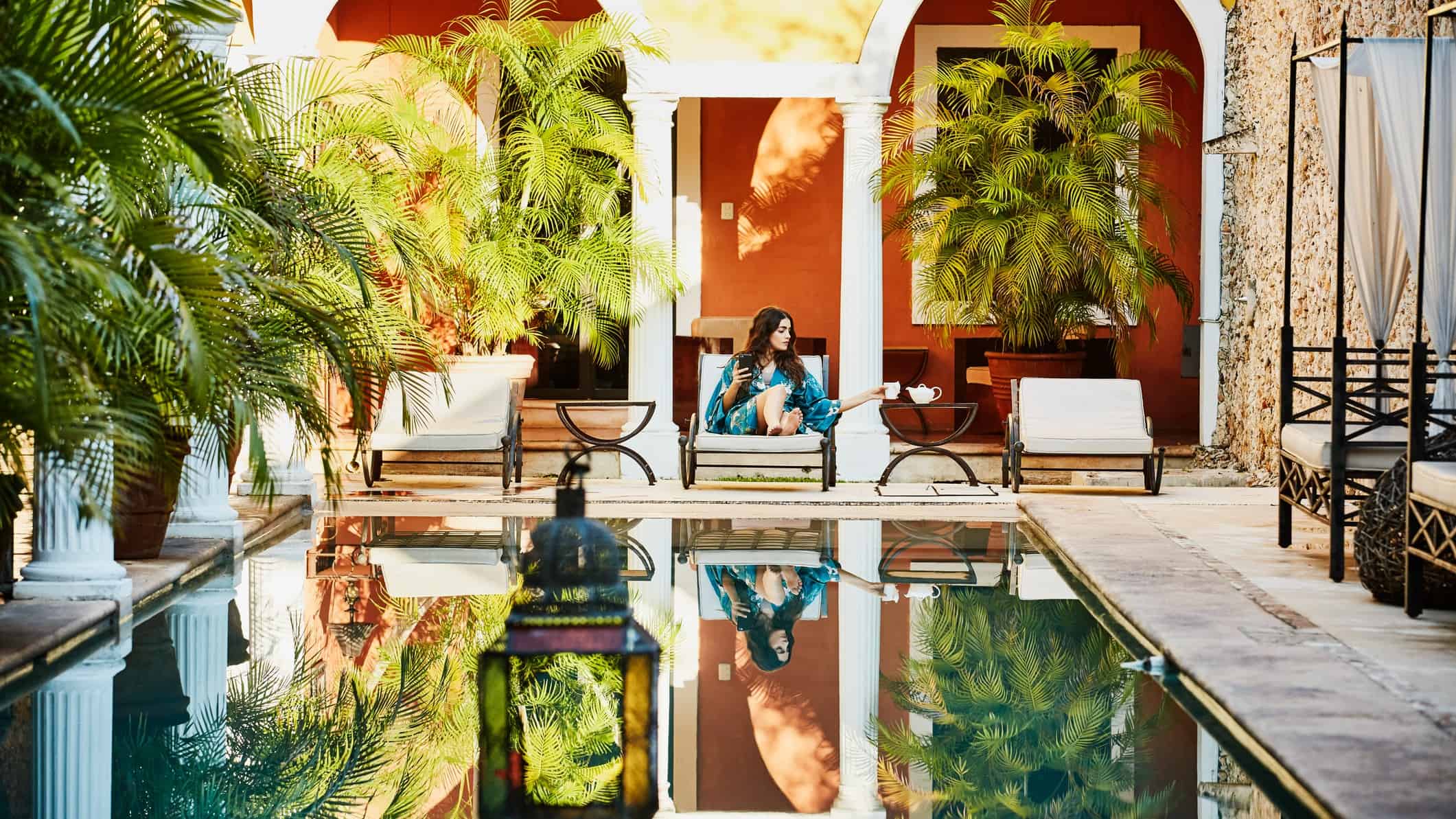 a woman in a luxurious poolside setting looks at her phone while drinking tea in a palace-style courtyard.