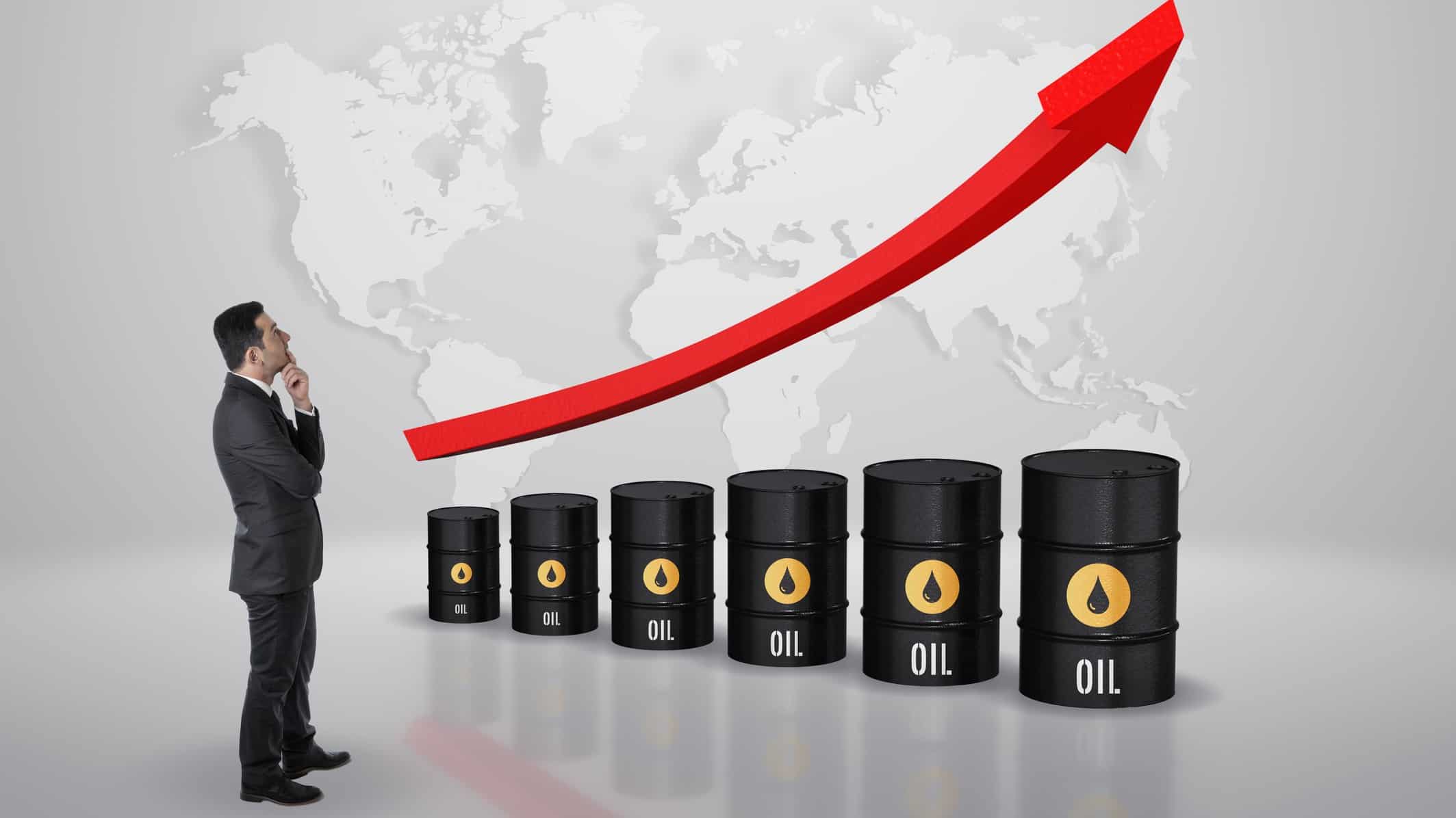 a man in a business suit looks at a map of the world above a line up of oil barrels with a red arrow heading upwards above them, indicting rising oil prices.