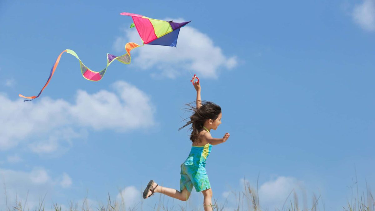 A girl runs along with her kite flying high in the sky.
