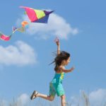 A girl runs along with her kite flying high in the sky.