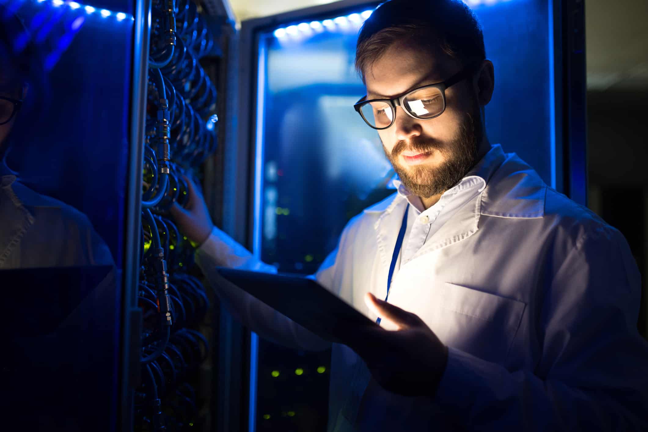 A young technician stands in a dark computer server room looking at his ipad during a cybersecurity inspection