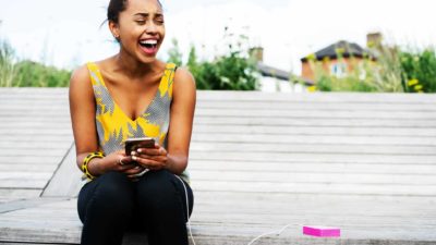 A woman sits on a step laughing at something on her mobile phone as it is being charged by a lithium-powered battery.