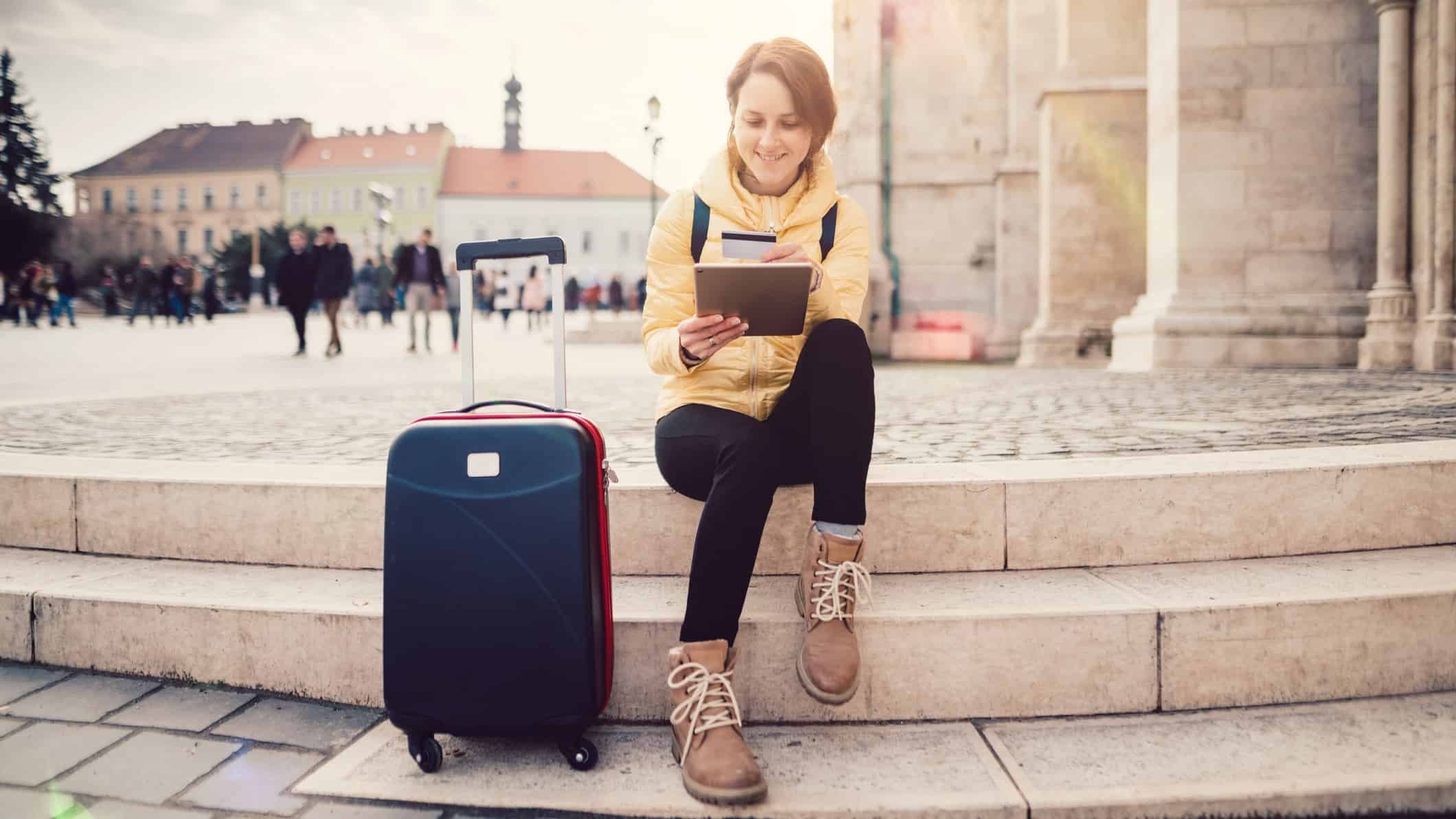 A young woman makes an online travel booking as she sits on some steps with her suitcase next to her.
