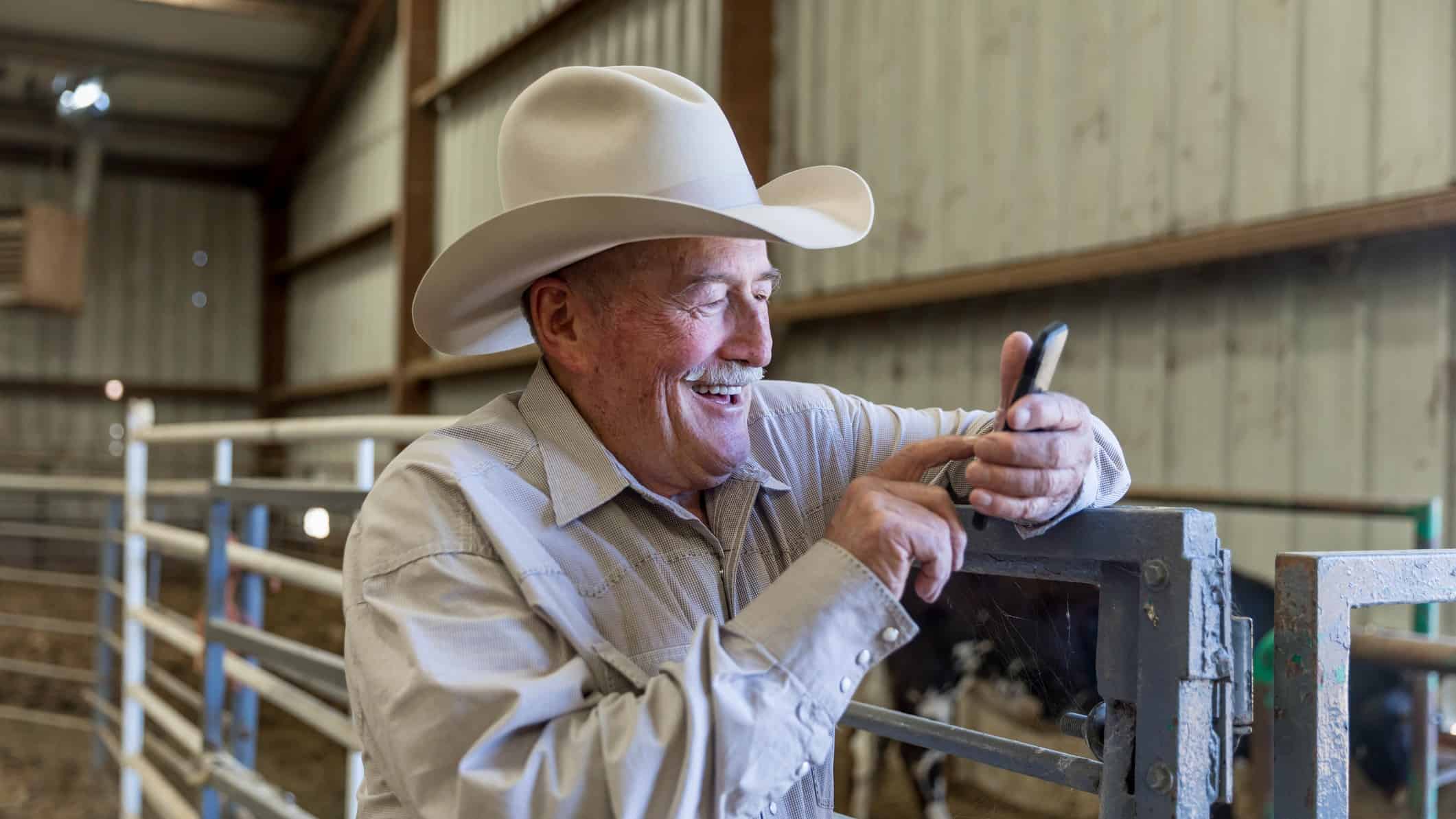 en äldre man i en cowboyhatt gör en handel på sin telefon medan han lutar sig mot häststallet.