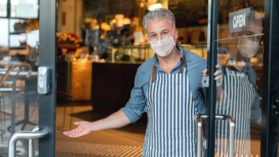 A shopkeeper wearing a mask reopens for business, holding his door open to welcome customers in.