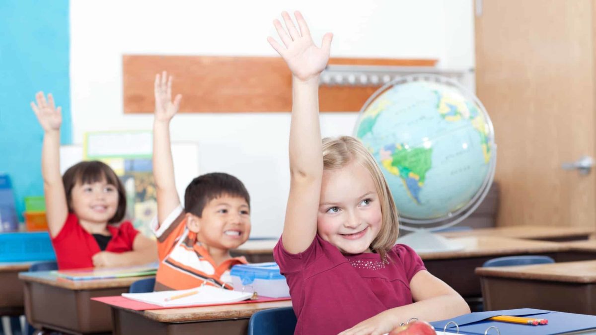 Three little kids in the classroom with their hands in the air, eager to answer a question.