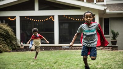 Two boys in capes running on the grass in front of their house.