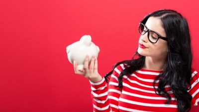 A woman dressed in red and standing in front of a red background peers thoughtfully at a piggy bank in her hand.