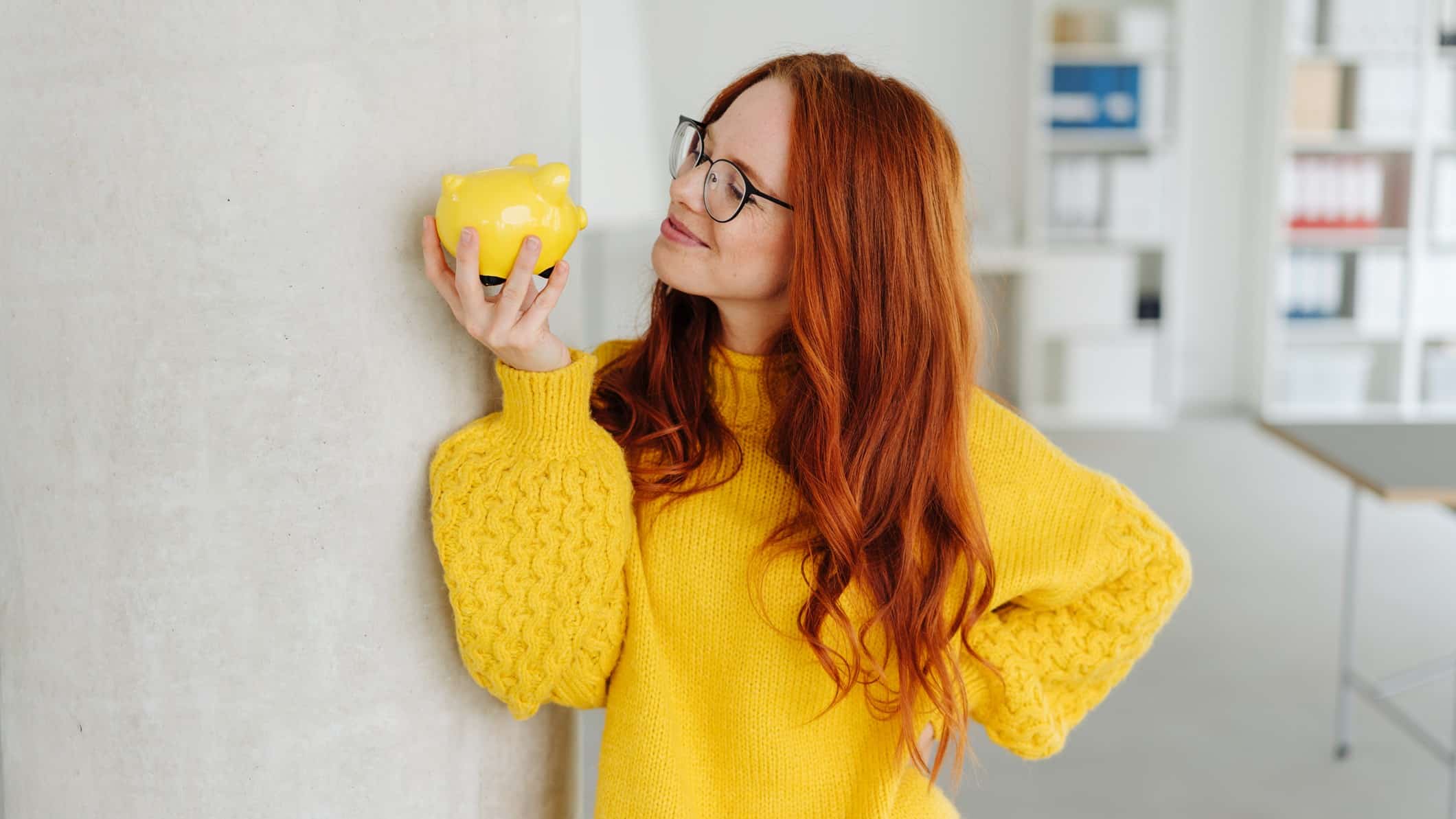 A woman in a bright yellow jumper looks happily at her yellow piggy bank.