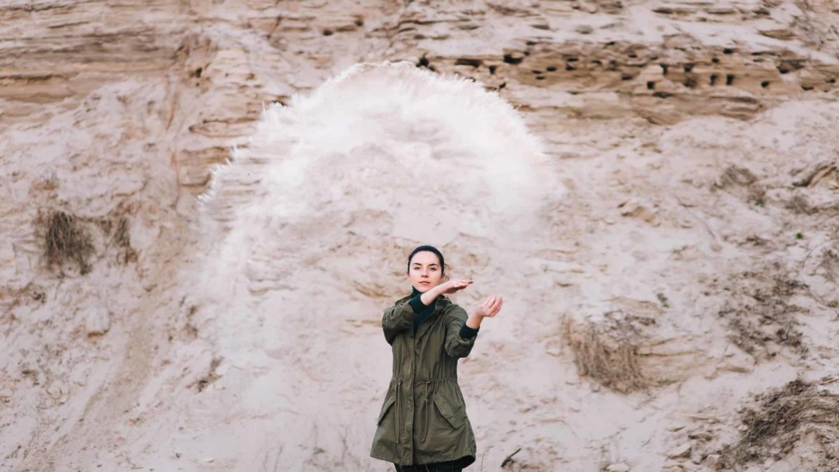 A statuesque woman throws earth in the air in front of a rocky outcrop.