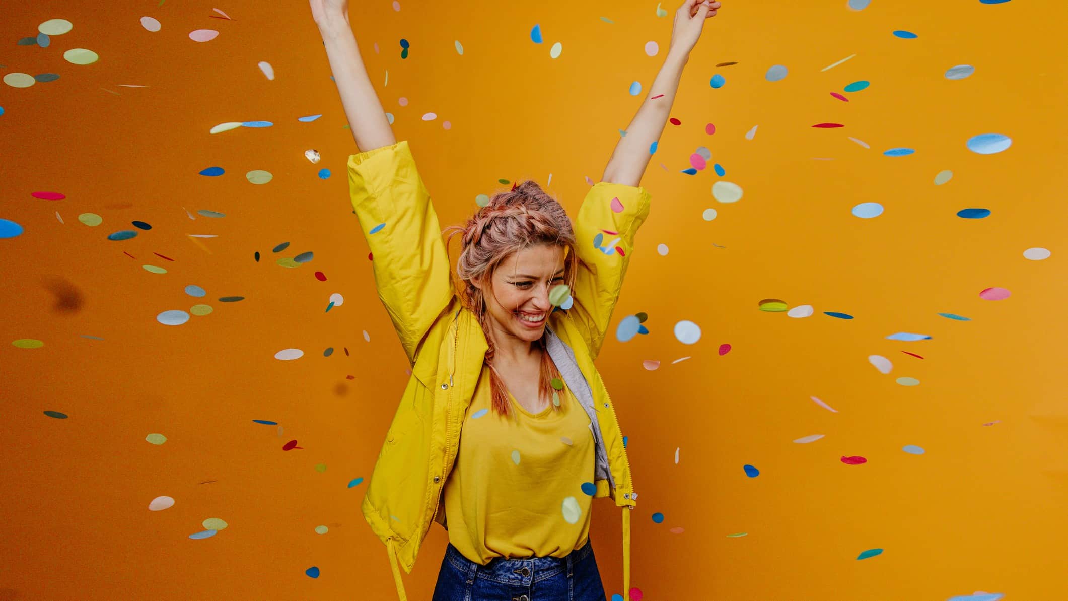 A woman throws her hands in the air in celebration as confetti floats down around her, standing in front of a deep yellow wall.