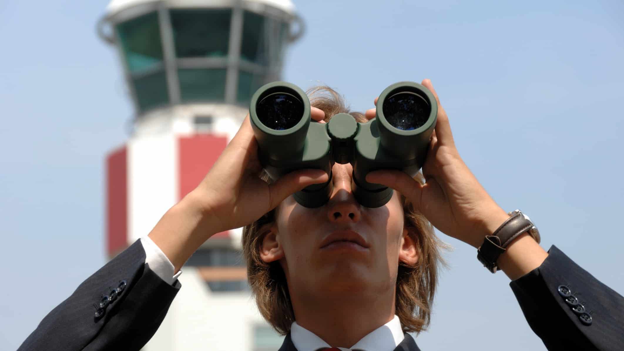 Man in suit looks through binoculars in front of a control tower at an airport.
