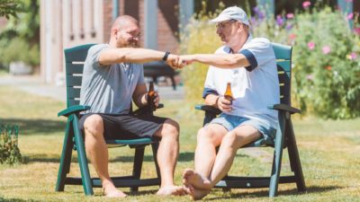 Two men sit in garden on chairs facing each other and fist bump while holding a beer.