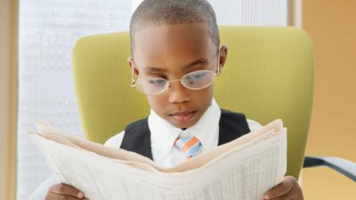 Young boy with glasses in a suit sits at a chair and reads a newspaper.