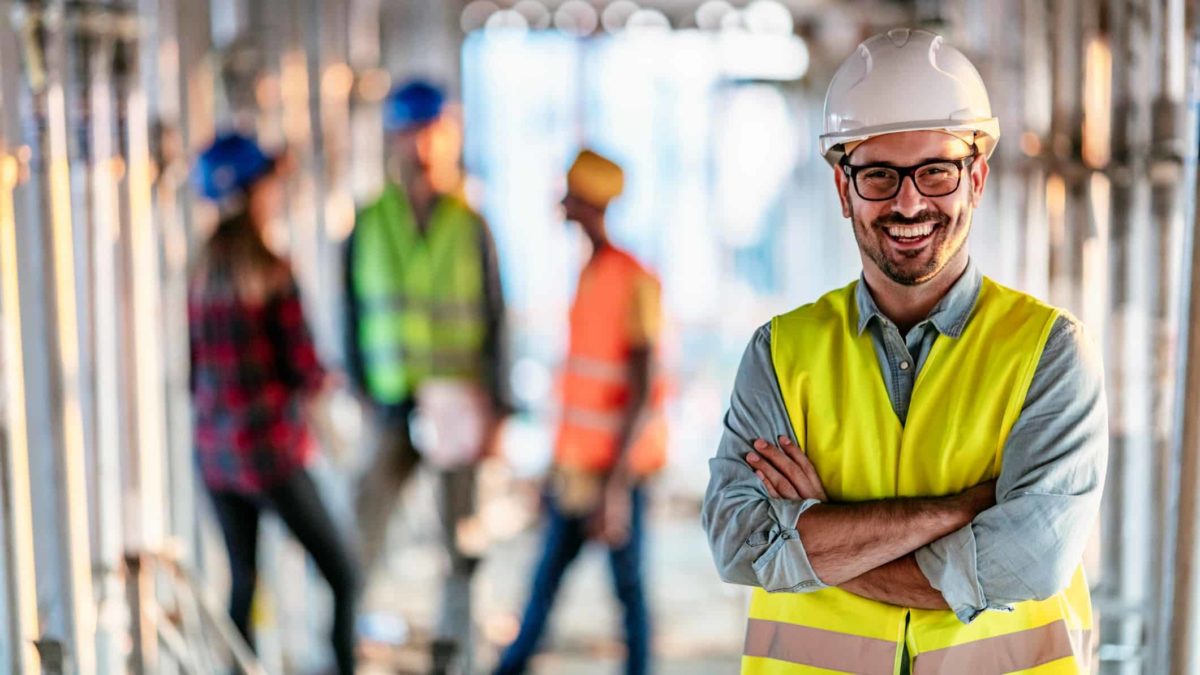 Male building supervisor wearing high vis vest and hard hat stands and smiles with his arms crossed at a building site