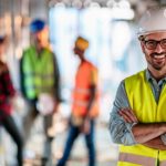 Male building supervisor stands and smiles with his arms crossed at a building site with workers behind him.