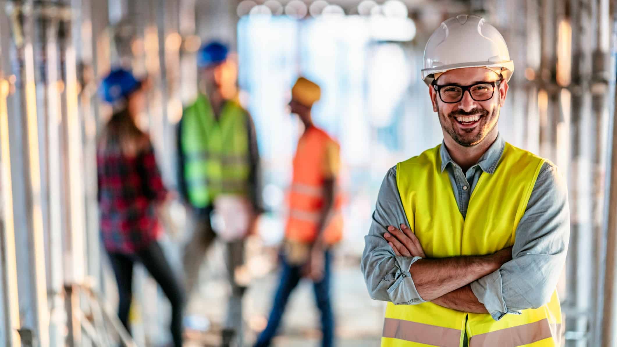 Male building supervisor stands and smiles with his arms crossed at a building site with workers behind him.