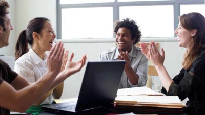 A group of four business people sit around a desk and laptops clapping and smiling.