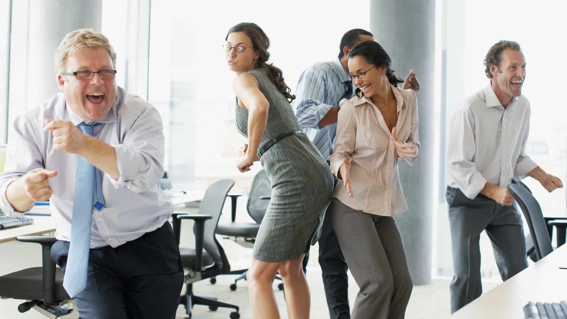 A group of business people dance around the office looking very happy.
