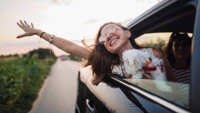A smiling woman with a cute dog flings her arm out of the window of a car