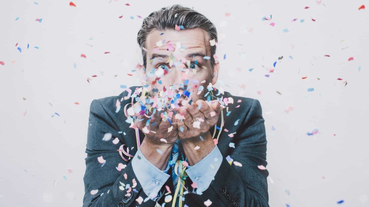 A man dressed in grey suit blowing confetti out of his hands towards the camera looking happy.