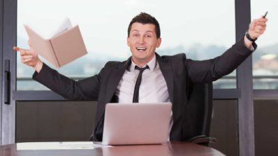 Man sitting at a laptop in an office throws a book into the air and cheers.