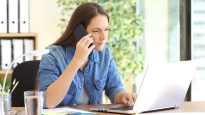 Woman looks puzzled as she types on laptop and uses phone.