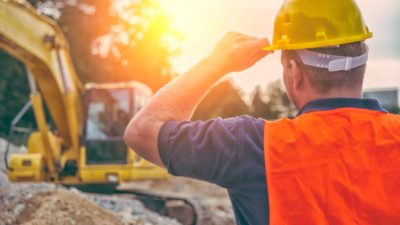 Builder with back to camera wearing hard hat watching tractor earthmover in sunset