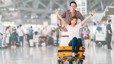 Man wheels trolley full of suitcases while woman sits on them with her hands in the air at an airport.