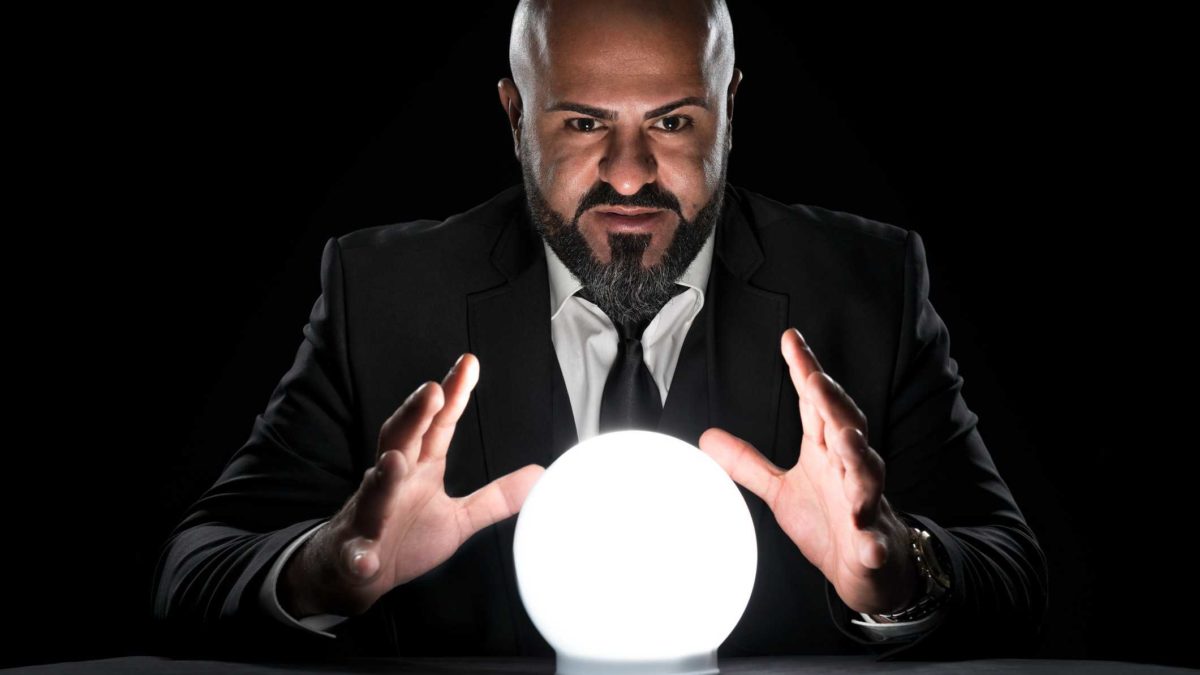 A bald man in a suit puts his hands around a crystal ball as though predictin the future.