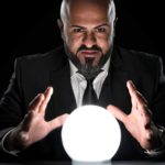 A bald man in a suit puts his hands around a crystal ball as though predicting the future.