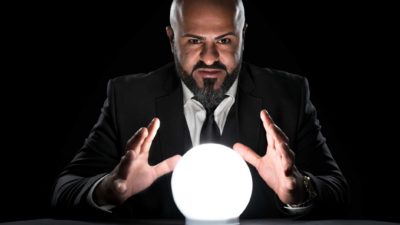 A bald man in a suit puts his hands around a crystal ball as though predicting the future.