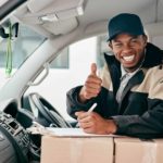 A delivery driver leans on boxes in his van as he puts his thumb up.