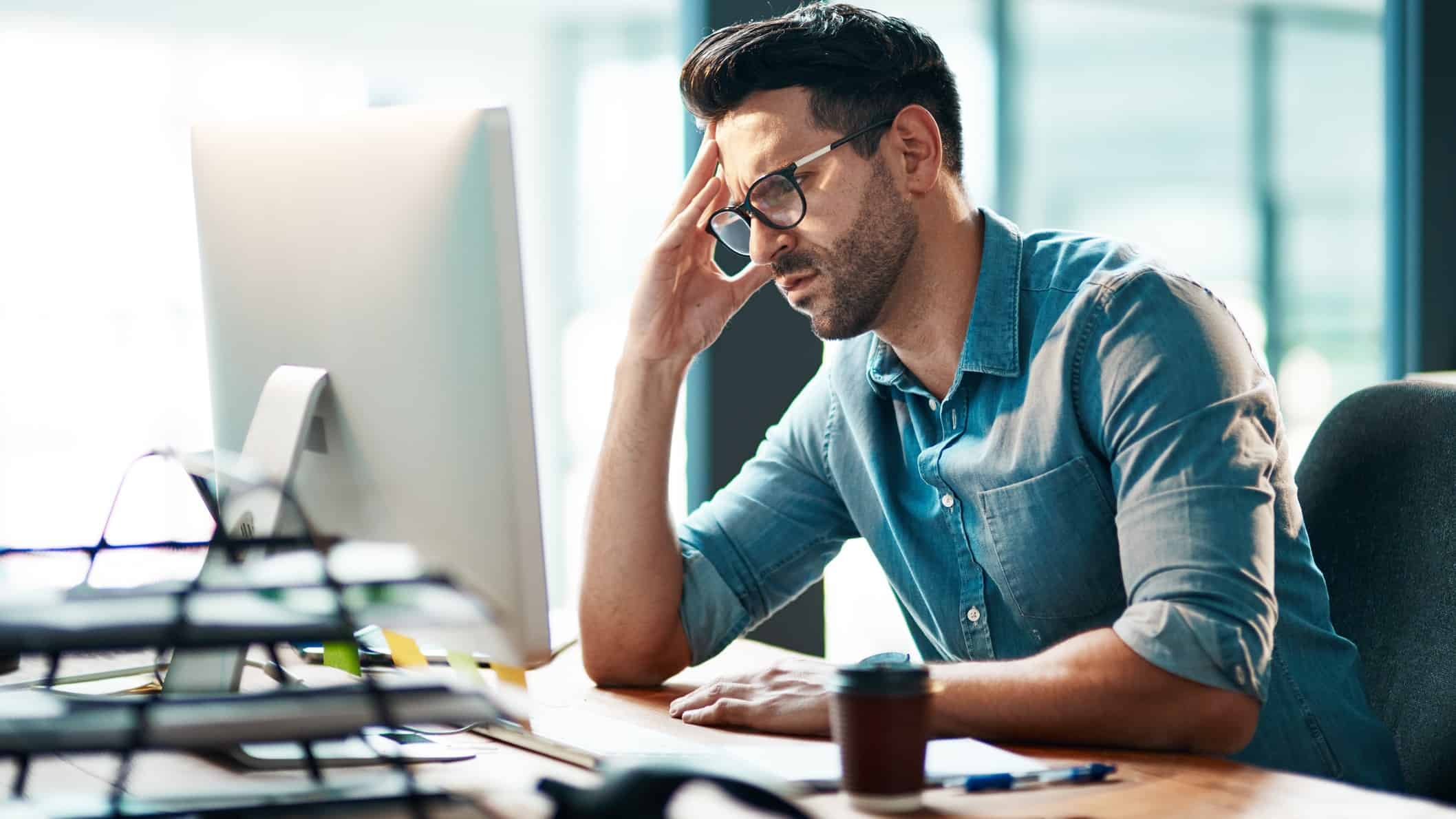 A male executive worker wearing glasses and a blue collared shirt looks at his laptop screen with a concerned look on his face and his hand to his forehead as he watches the Bank of Queensland share price fall