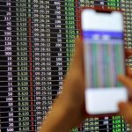 Person holding up a smartphone in front of a stock market chart.