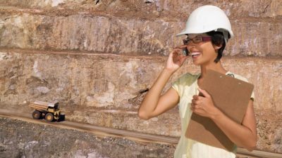 A mining executive from Red Dirt Metals chats on her mobile phone looking pleased with a mining site and mining truck in the background