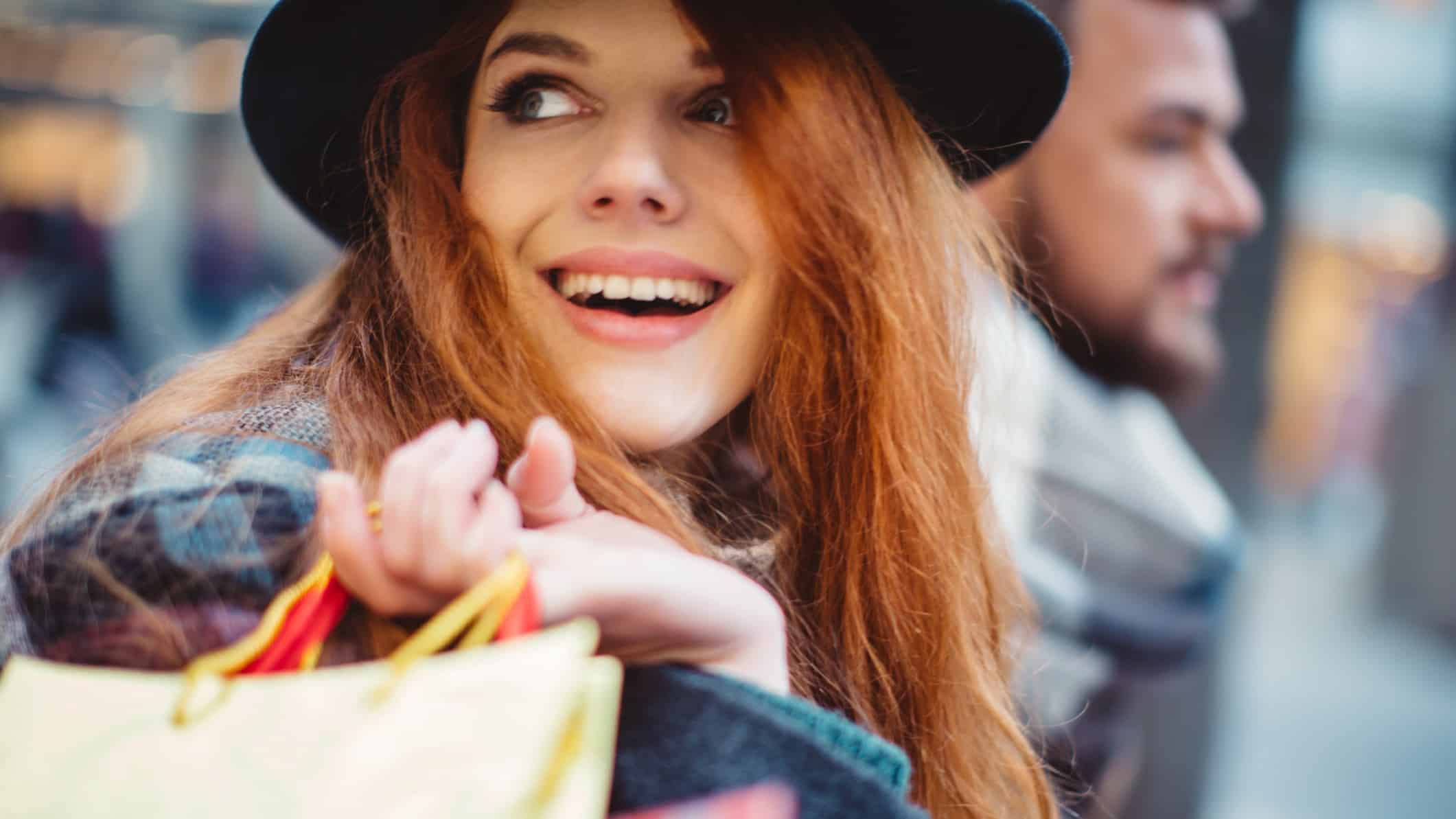 Close-up of a woman waring a hay and smiling as she carries shopping bags over her shoulder.